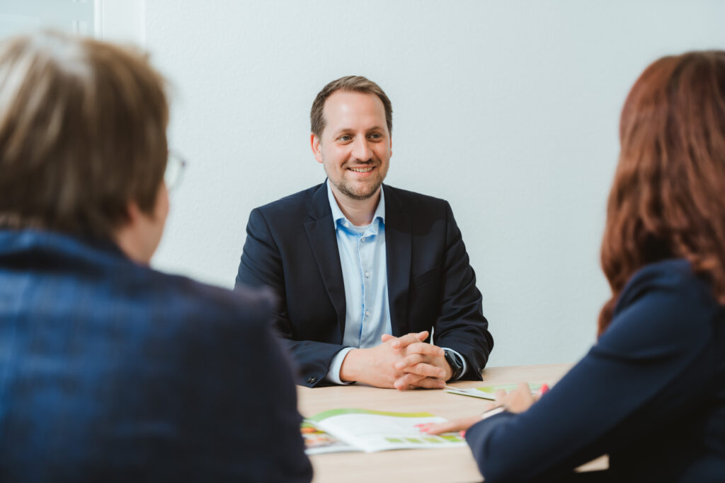 Foto von Jan-Eric Hennig. Er sitzt, wie in einem Beratungsgespräch, an einem Tisch. Gegenüber von ihm sitzen Joanna Tusche und Heike Horstmann. Vor ihnen liegen Prospekte der LVM Versicherung.