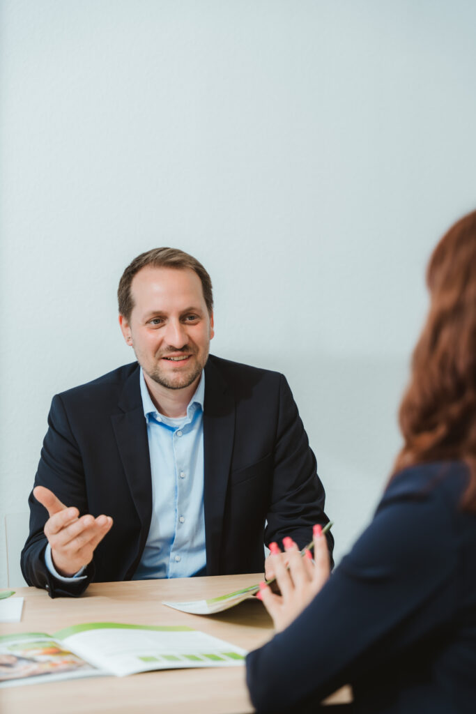 Foto von Jan-Eric Hennig. Er schaut freundlich, gestikuliert leicht und sitzt an einem Tisch. Gegenüber von ihm sitzt Joanna Tusche. Vor den beiden liegen Prospekte der LVM Versicherung.