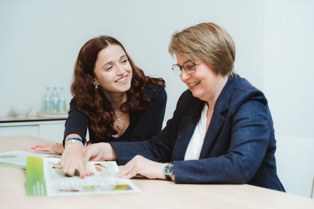 Foto von Joanna Tusche und Heike Horstmann. Sie sitzen an einem Tisch und schauen gemeinsam in Projekte der LVM Versicherung.